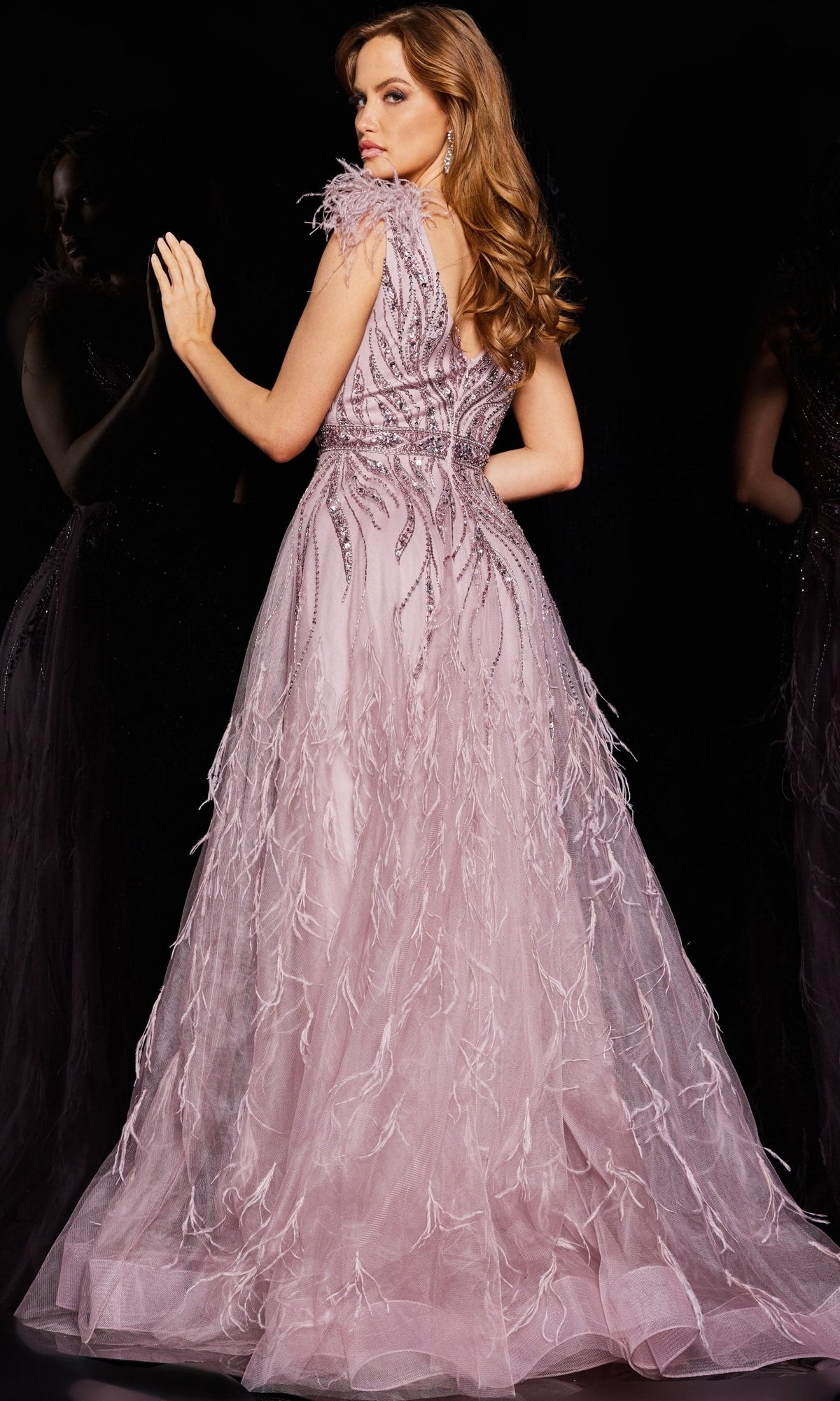 A woman stands against a black background, facing away, wearing the Jovani Feathered Long Pink Prom Dress 24000—an elegant beaded gown with feather-like embellishments.